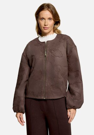 Femme portant une veste marron zippée avec broderie florale et manches bouffantes, associée à un pantalon marron foncé et un haut blanc à col volanté.
