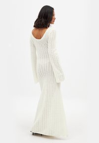 Robe longue blanche en tricot avec un dos dégagé, des manches longues et une silhouette ajustée, présentant un motif texturé à mailles ouvertes.