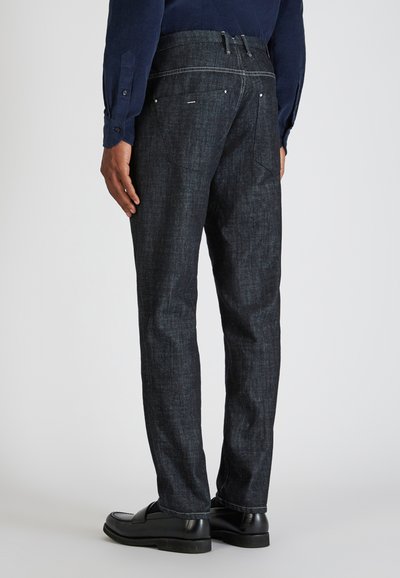 Mann trägt dunkelblaue Leinenhosen, schwarze Lederloafers und ein navyblaues Langarmshirt, steht mit dem Rücken zur Kamera.