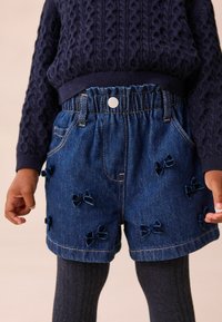 Denim shorts med elastisk talje, blå sløjfer syet på stoffet, parret med mørke strikketights og en navy struktureret sweater.