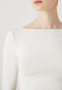 Top alb cu maneci lungi și decolteu barco. Material neted, ușor, cu un ușor luciu, design ajustat, detalii de cusătură minime.