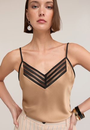 Femme portant un haut beige sans manches avec un col en maille rayée noire, un pantalon beige rayé, des boucles d'oreilles en perles et des bracelets noirs et dorés.