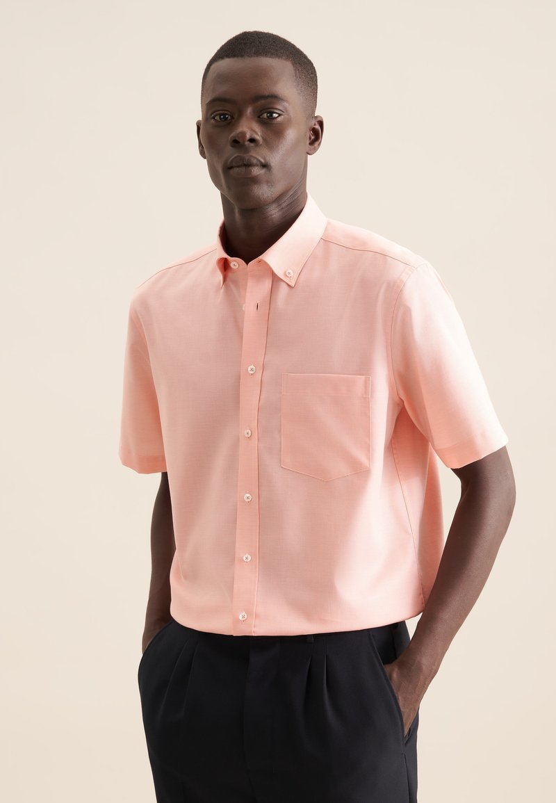 Camicia a maniche corte in tessuto rosa chiaro, con colletto a punta, taschino sul petto e bottoni bianchi. Indossata con pantaloni scuri.