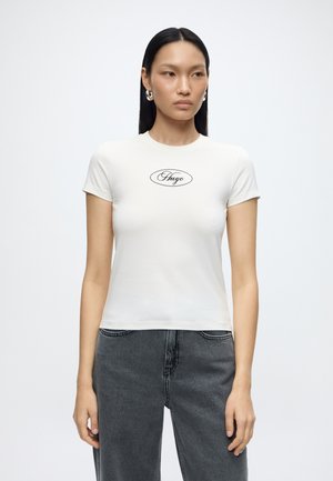 Femme aux cheveux noirs raides portant des boucles d’oreilles argentées en anneau, un T-shirt blanc ajusté avec le logo « Hugo » et un jean gris foncé, debout sur fond blanc.
