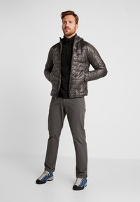 Puffige graue Jacke mit Kapuze, strukturierter Stoff, Reißverschluss; getragen über einem schwarzen Oberteil, grauen Hosen und blauen Wanderschuhen mit weißen Schnürsenkeln.