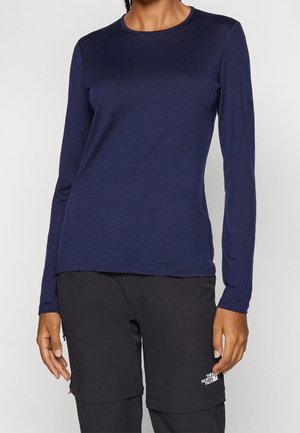 Personne portant un t-shirt bleu marine à manches longues et un pantalon d'extérieur noir The North Face, debout les bras détendus.