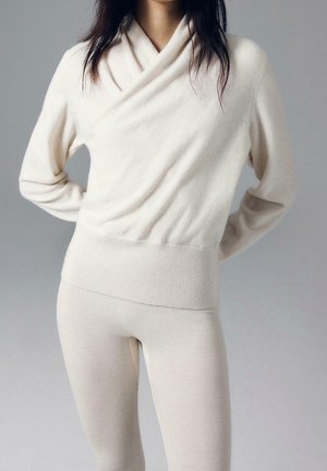 Femme portant un pull en maille couleur crème avec un col croisé et un pantalon en maille ajusté assorti, sur un fond uni.