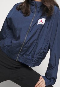 Granatowa, lekka kurtka wykonana z błyszczącego materiału, z wysokim kołnierzem, zamkiem błyskawicznym oraz białą łatą z logo i czerwonymi akcentami.
