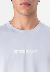 Světlé šedé tričko s kulatým výstřihem vyrobené z bavlny, s vyšitým logem "GUESS JEANS" v bílé barvě, krátkými rukávy a hladkou texturou.