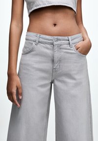 Gros plan sur le buste d'une personne portant un jean gris clair taille haute à jambes larges et un crop top, une main dans la poche et l'autre détendue le long du corps.