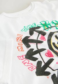 T-shirt blanc avec des flèches noires formant un cercle, des cœurs rouges, des fleurs vertes et un texte coloré disant "Ose," "Temps," et "Habite."