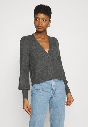 Cardigan grigio lavorato a maglia con scollatura a V profonda, maniche lunghe a sbuffo e cinque bottoni sul davanti. Abbinato a jeans in denim blu chiaro.