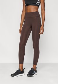 Leggings marron à coupe ajustée avec une taille haute, confectionnés dans un matériau extensible. Associés à des chaussures de sport noires avec des accents blancs.