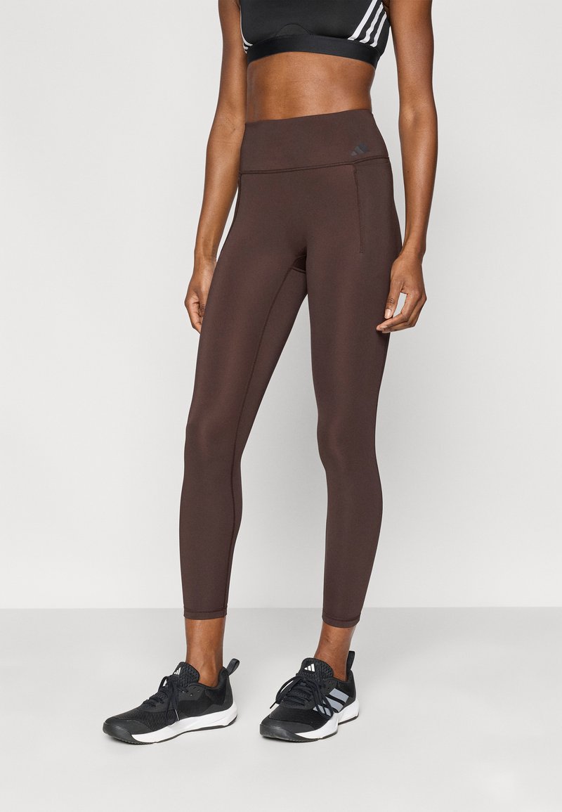 Leggings marron à coupe ajustée avec une taille haute, confectionnés dans un matériau extensible. Associés à des chaussures de sport noires avec des accents blancs.