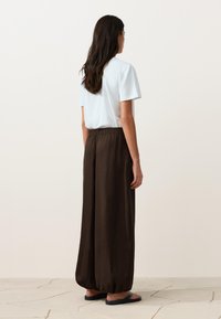 Femme aux longs cheveux foncés portant un haut blanc à manches courtes, un pantalon large ample marron foncé et des sandales noires, debout sur un sol carrelé.