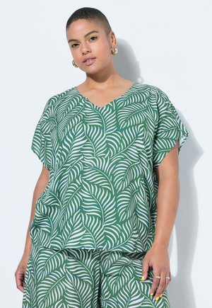 Frau mit kurzen Haaren, die ein grünes und weißes, mit Blättern gemustertes Oberteil und passende Hosen trägt, goldene Creolen und gelben Lidschatten.