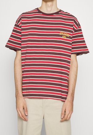 Osoba ubrana w T-shirt z poziomymi paskami w kolorach czerwonym, białym i brązowym z logo "BDG JEANS" na piersi, w połączeniu z beżowymi spodniami.