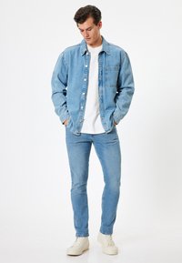 Camicetta in denim azzurro chiaro con taschino, indossata sopra una t-shirt bianca, abbinata a jeans azzurri chiari e sneakers bianche. Texture liscia, vestibilità casual.