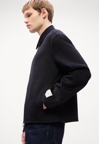 Veste noire texturée avec un col, des poches latérales et un accent de label blanc sur la manche ; portée sur un jean en denim, vue de profil.