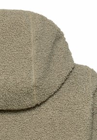 Zbliżenie na beżowy, teksturowany kaptur z polaru i szew na ramieniu miękkiej, ciepłej kurtki lub płaszcza.