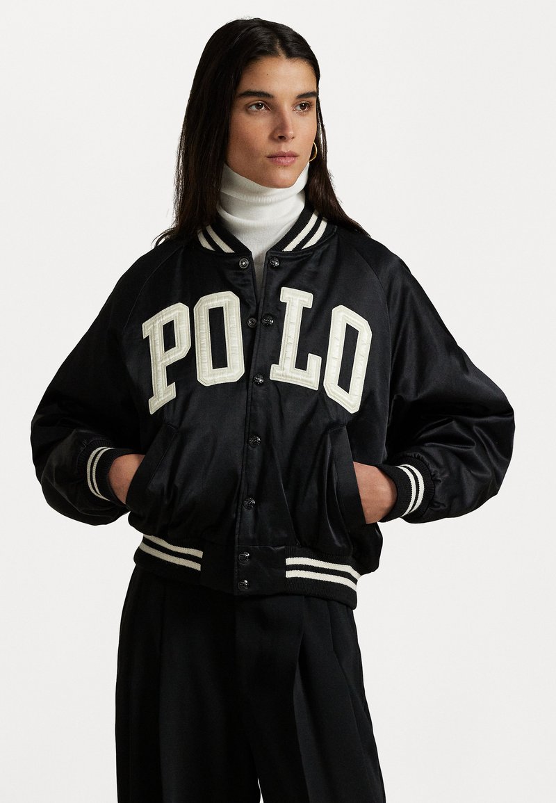 Must mustri bomber-jakk must valge "POLO" kirja, triibuline ribikätikud ja äär, nööpkinnis, ning küljetaskud.