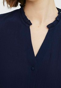Blouse bleu marine en tissu texturé, avec un col à volants et une fermeture à bouton à l'avant. Design en V.