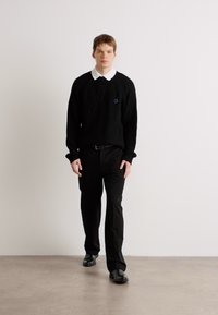 Pulover negru din tricot cu guler alb, asortat cu pantaloni și pantofi negri. Puloverul are un mic logo albastru pe piept.