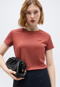 Νεαρή γυναίκα με κοντά κυματιστά μαλλιά φοράει ένα μπλουζάκι BOSS σε σκουρόχρωμη κεραμιδί απόχρωση και κρατά μια μαύρη τσάντα φάκελο από δέρμα, σε απλό φόντο.