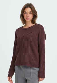 Pull en tricot bordeaux avec un col rond, texture côtelée et manches longues. Détails de coutures diagonales remarquables sur le corps.