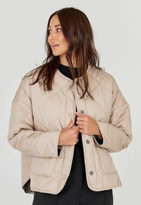 Heledbeez värvi tikitud jakk ümmarguse kaelusega, nööpsulguritega ja küljetaskutega. Kangas näeb välja siledana ja on veidi polsterdatud.