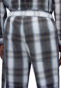 Vue arrière d'une personne portant un pantalon à carreaux gris et bleu clair et un haut assorti, avec les mains partiellement visibles sur les côtés.