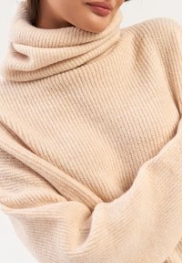 Beżowy sweter z golfem w prążki o miękkiej teksturze, z zagiętym kołnierzem i luźnymi rękawami.