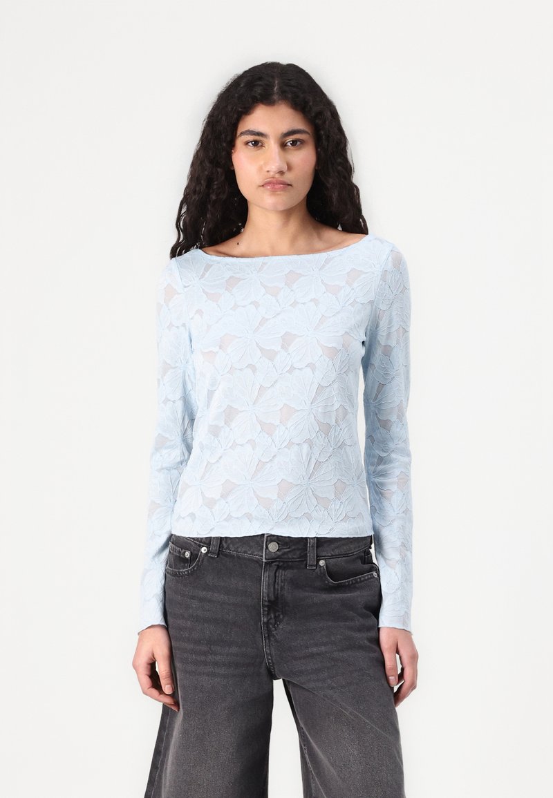 Light blue long-sleeve top with floral lace design, sheer texture, wide neckline, paired with dark gray high-waisted jeans.
