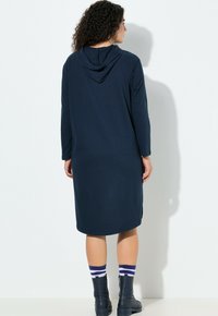 Długa granatowa sukienka z kapturem z długimi rękawami, bocznymi rozcięciami, miękką fakturą i kieszenią kangurka. Noszona z niebieskimi botkami i paskowanymi skarpetkami.