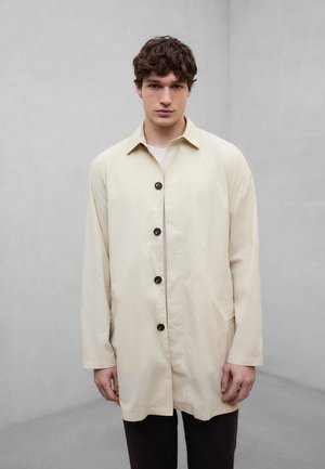 Ung mann med kort brunt hår, iført en lys beige knappet frakk og mørke bukser, står mot en ensfarget grå bakgrunn.