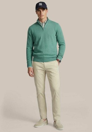 Mann trägt dunkelblauen Baseballcap, grünen Pullover mit Viertelreißverschluss, gestreiftes Hemd, beige Hose, beige Mokassins und Armbanduhr, steht vor einfarbigem Hintergrund.