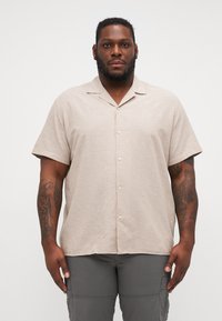 Camicia beige a maniche corte con bottoni, realizzata in tessuto leggero, con colletto e una leggera texture, indossata con pantaloni cargo grigi.