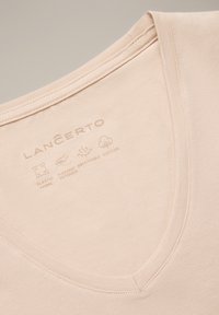 Beżowa koszulka z dekoltem w serek wykonana z elastycznej, oddychającej bawełny. Napis na dekolcie informuje o właściwościach tkaniny: miękka, elastyczna i oddychająca.