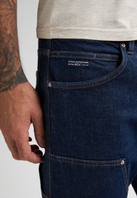 Jeans in denim blu scuro con un design aderente, caratterizzati da cuciture arancioni, tasche frontali e un'etichetta del marchio sulla tasca posteriore.