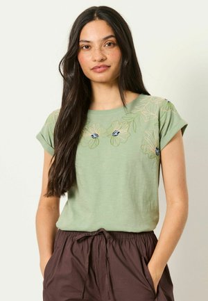 Young woman with long dark hair wears a light green floral embroidered shirt and dark brown drawstring pants, hands in pockets.