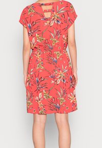 Blomstret kjole i koralfarvet stof, med korte ærmer, en nøglehul i ryggen og en elastisk talje med løs pasform. Mønstret indeholder forskellige blomster.