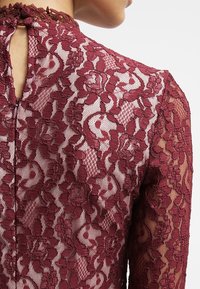 Haut en dentelle bordeaux avec des motifs floraux, manches longues et une fermeture au dos en forme de goutte. Tissu texturé avec une superposition transparente.