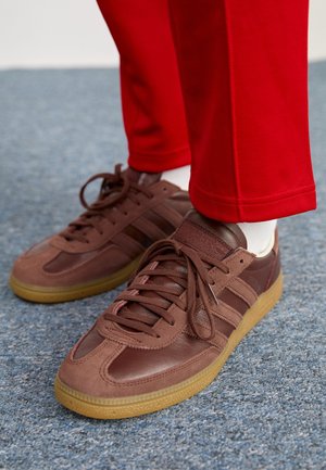 Baskets en cuir marron avec semelles en gomme portées avec des chaussettes blanches et un pantalon rouge sur une moquette bleue.