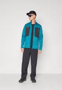Miękka kurtka w kolorze teal z czarnymi łatami, zamkiem błyskawicznym i logo. Połączona z czarnymi spodniami i butami. Model nosi okulary przeciwsłoneczne i czapkę.