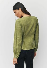 Oliivinvihreä, teksturoitu pusero, jossa pitkät hihat, röyhelöity kangas ja hieman levenevä helma, yhdistetty tummiin farkkuihin.