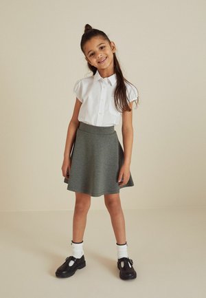 Hymyilevä nuori tyttö, jolla on pitkät hiukset, valkoinen lyhythihainen paita, harmaa hame, valkoiset sukat ja mustat remminilkkakengät, seisoo neutraalilla taustalla.