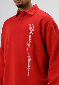 Rode polo met lange mouwen van katoen, voorzien van een kraag en geborduurde tekst "Young Atelier" in het wit langs de zijkant.