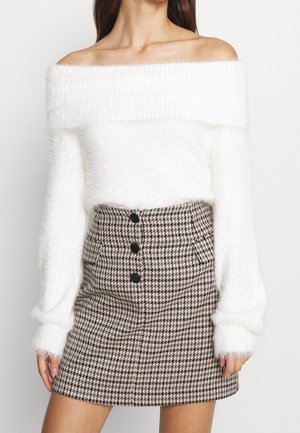 Femme portant un pull blanc duveteux à épaules dénudées associé à une jupe taille haute à motif pied-de-poule noir et blanc avec des boutons sur le devant.