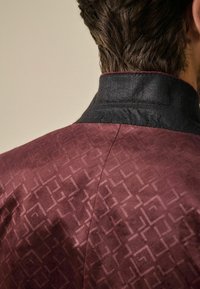 Jachetă maroon cu un model geometric texturat, având un guler negru și un material neted și moale. Cusături vizibile pe cusătura umărului.