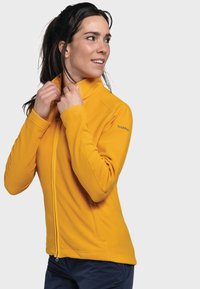 Gelbe Fleecejacke mit hohem Kragen, durchgehendem Reißverschluss und seitlichen Taschen. Glatte Textur mit genähten Nähten und Logodetail an der Schulter.